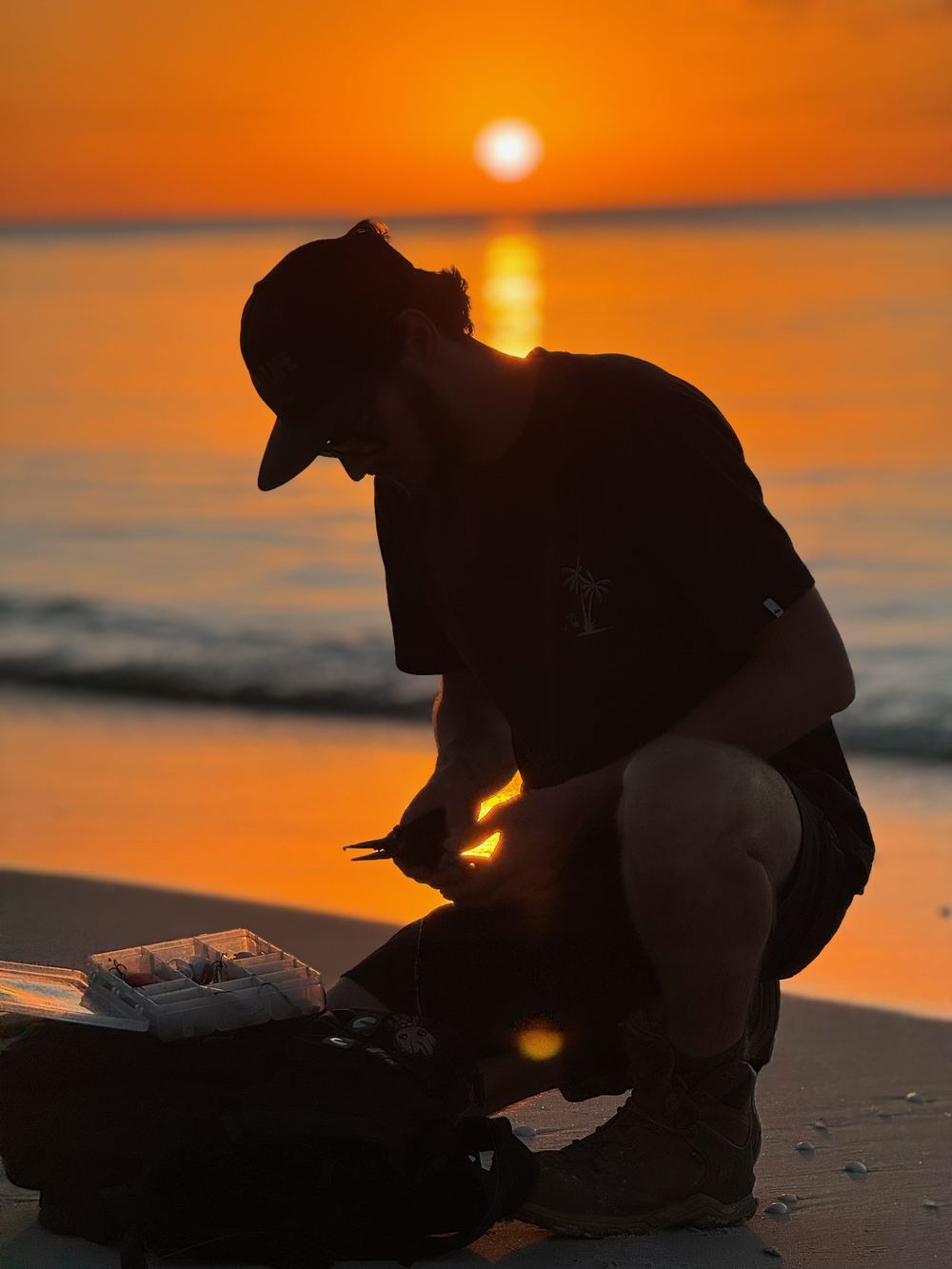 An angler in silhouette crouches by an open tackle box on the beach, rigging up as the sun sets over the water.