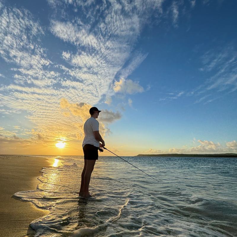 An angler wading in shallow surf casts towards the sunset.