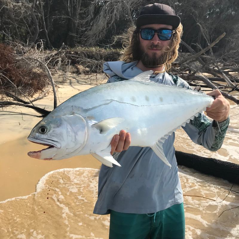 An angler in a cap and sunglasses holds up a big silver fish on the beach.