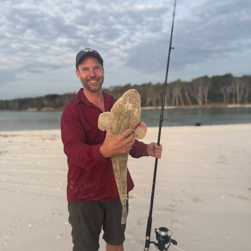 An angler on a sandy beach holds up a fish beside a surf rod.