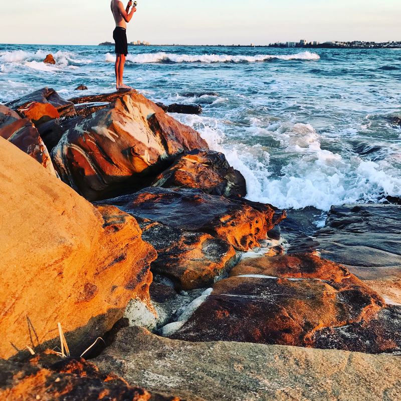 An angler stands on orange rocks above breaking waves, rod in hand.